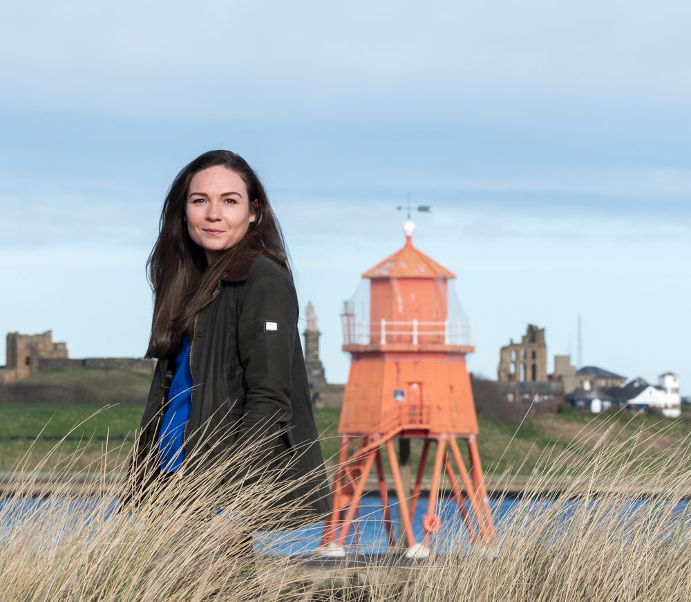 Georgie standing in the dune grass by the river mouth, with a small lighthouse behind her